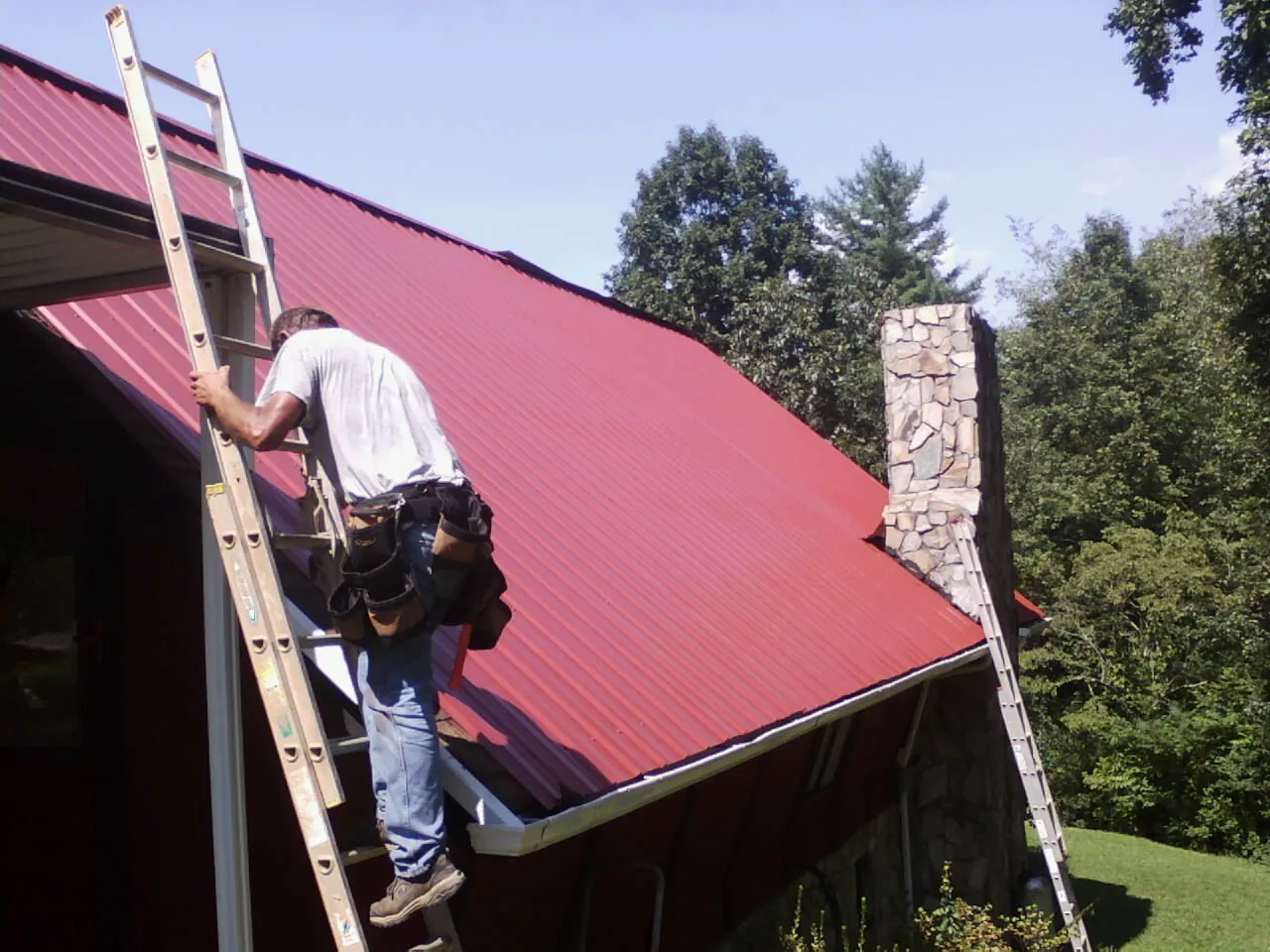 Roofer on a ladder beside a red metal roof and stone chimney — Soffit & Fascia Repair by Roofing Brothers in Sylvan Beach